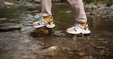 Yakın çekim: beyaz spor ayakkabılı bir kız taştan yapılmış özel bir patika boyunca bir dağ nehrinden geçiyor, nehrin diğer tarafında duran erkek arkadaşı ona yardım ediyor..