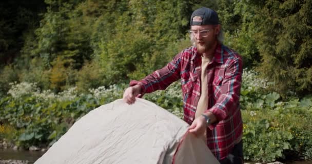一个戴着红色格子花衬衫 留着胡子的金发男人在旁边开枪 在森林的背景下搭起了一个帐篷 — 图库视频影像