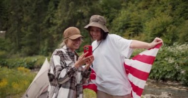 Şapkalı sarışın bir kız, üzerinde Amerikan bayrağı olan beyaz tişörtlü esmer bir kızı gösteriyor. Ormanda yürüyüş yaparken çekilmiş bir fotoğraf..