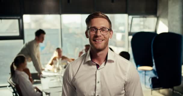 メガネのブロンドの男とオフィスの白いシャツは パノラマの窓のあるオフィスのテーブルで働く彼のゴールデンレグの背景に対してポーズします — ストック動画