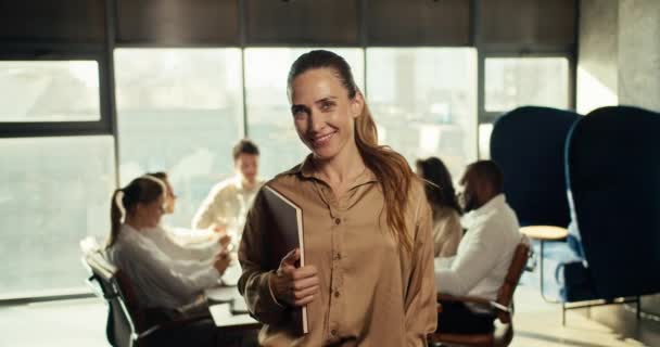 穿着棕色商业服装的成功的女商人微笑着看着办公室里的摄像机 在办公室工作人员的背景下 — 图库视频影像