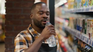 Ekose gömlekli siyah tenli bir adam süpermarketteki şeffaf plastik bir şişeden su içer..