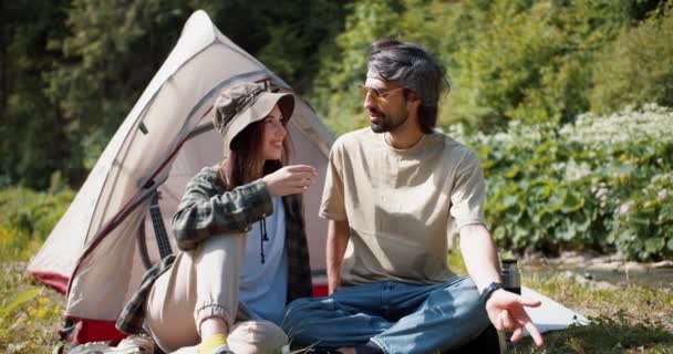 男孩和女孩坐在帐篷旁边聊天 女孩喝着热水瓶里的茶 森林背景下帐篷附近的定居点 — 图库视频影像