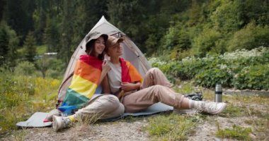 Sarışın bir kız ve esmer bir lezbiyen kız bir lgbt bayrağına sarılmış yeşil bir ormanın arka planında yan yana oturuyorlar..