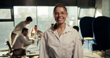 Gözlüklü mutlu bir kızın portresi ve ofiste gülümseyen ve kameraya bakan beyaz bir gömlek. Ofis çalışanlarının çalışma sürecinin arka planına karşı poz vermek.