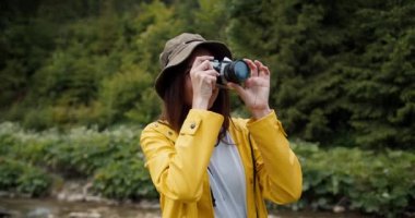 Sarı ceketli esmer bir kız bir ormanın ve bir dağ nehrinin fotoğrafını çekiyor..