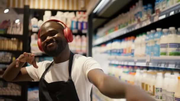 赤いヘッドフォンに黒い肌の色の男と スーパーマーケットの酪農部で 黒いエプロンが踊る 仕事で幸せに — ストック動画