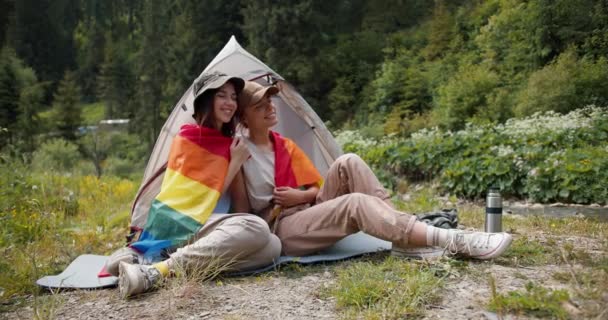 一个金发碧眼的女孩和一个黑发的女同志坐在一起 裹着一面大旗子 坐在一座绿色森林的背景下的一个帐篷旁边 — 图库视频影像