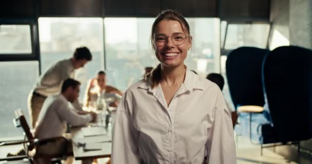 一个戴眼镜的快乐女孩的画像和办公室里的一件白色衬衫 她微笑着看着相机 在办公室工作人员工作进程的背景下提出的建议 — 图库视频影像