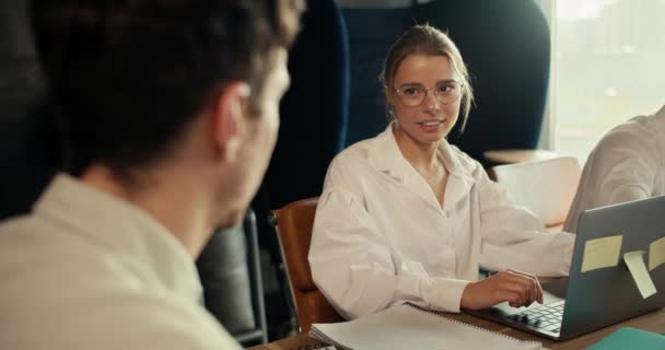 穿着白衬衫戴着眼镜的金发女孩与穿着白衬衫的员工交流 她们坐在办公室的桌子旁 桌上有笔记本电脑 — 图库视频影像