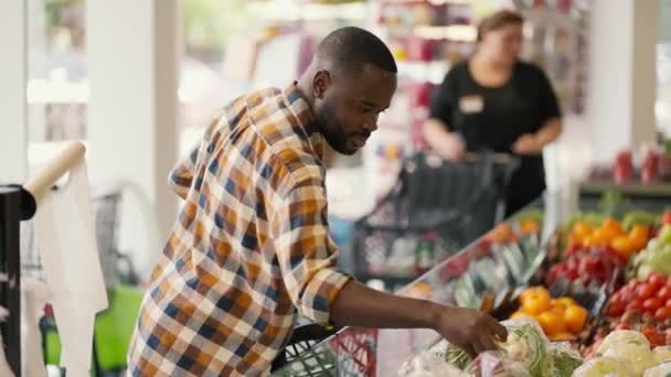 一位穿着格子呢衬衫的黑皮肤男子在超市柜台上挑选水果和蔬菜 男人闻到货物的味道 就会选择他需要的 — 图库视频影像