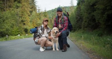 Yürüyüş kıyafetleri içinde bir adam ve bir kız, açık renkli köpekleri ile birlikte, poz verir ve ormanın arka planına karşı yolun kenarında dururken kameraya bakarlar..