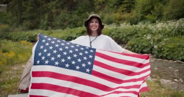 Beyaz tişörtlü, vatansever esmer bir kızın portresi. Amerika Birleşik Devletleri bayrağıyla birlikte yeşil bir ormanın ve bir dağ nehrinin zeminine gülümsüyor..