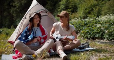 Esmer bir kız Amerika Birleşik Devletleri bayrağına sarılmış ve kamp kıyafetleri giymiş bir kız gitar çalıyor, yeşil bir ormanın arka planına karşı çadırın yanında oturuyorlar..