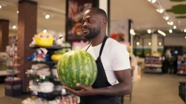 Beyaz tişörtlü ve siyah önlüklü siyah bir adam bir karpuz taşıyor ve süpermarket tezgahına koyuyor. Yaz mevsiminde süpermarket.