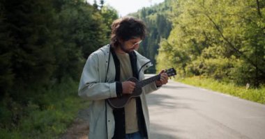 Yol kenarında beyaz ceketli esmer bir adam dağlarda bir yürüyüş yaparken müzik aleti çalıyor..