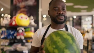 Siyah derili beyaz tişörtlü ve siyah önlüklü bir adam bir karpuz taşıyor ve süpermarket tezgahına koyuyor. Yüksek kalitede çekilmiş video /