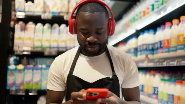 赤いヘッドフォンの黒い肌の色と赤いスマートフォンを持つ黒いエプロンを持つ男は スーパーマーケットの乳製品部門に沿って歩きます — ストック動画