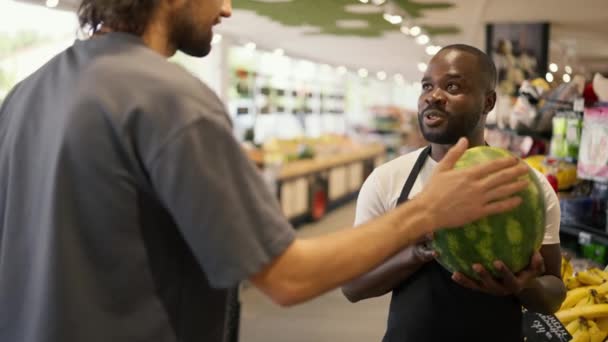一个黑皮肤的男人 穿着黑色围裙 手里拿着西瓜 谈论它 一位商店的顾客向一位顾问询问西瓜的事 — 图库视频影像