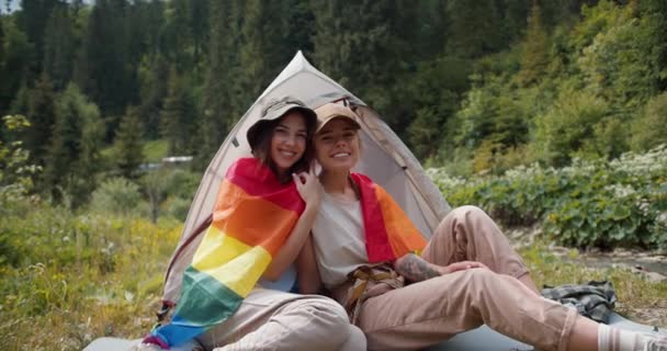 Lgbtフラッグに身を包んだ2人のレズビアン少女の肖像画は 緑の森の背景にテントの近くに座っている 非伝統的な性的指向を持つ人々のキャンペーン — ストック動画