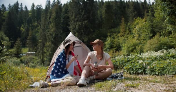 一个金发碧眼的姑娘和一个戴帽子的黑发姑娘 在美国国旗下游泳 坐在帐篷旁边的绿林中 与人交流 — 图库视频影像