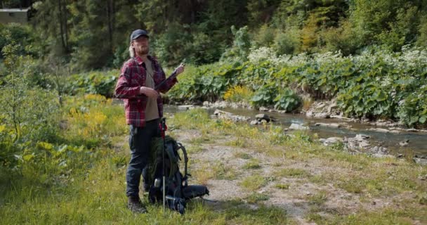 一位金发碧眼的游客 身穿红色登山衫 留着胡子 打开两根杆子 在森林小河边的草坪上散步 — 图库视频影像