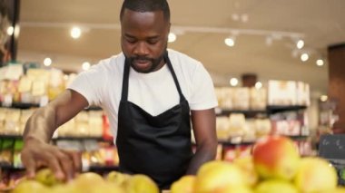 Yakın plan: Beyaz tişörtlü ve siyah önlüklü siyah bir adam süpermarket tezgahında sarı armutlar düzenliyor.