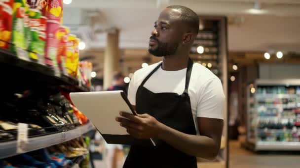 身穿白色T恤和黑色围裙的黑皮肤男子穿过超市的橱窗 用纸和平板电脑清点存货 — 图库视频影像
