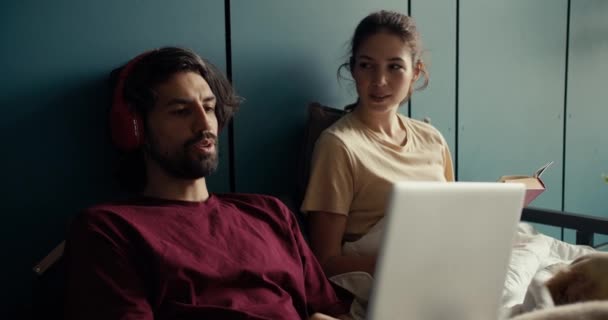 一个穿着红色T恤无线耳机的黑发男人在家里通过视频通信进行交流 在他的女朋友旁边坐着看书 一对快乐的夫妇在家里 — 图库视频影像