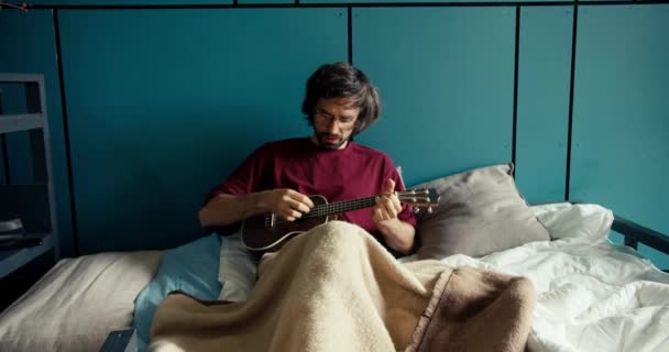 Ένας Μελαχροινός Τύπος Μούσι Κόκκινο Πουκάμισο Γυαλιά Παίζει Κιθάρα Ξαπλωμένος — Αρχείο Βίντεο