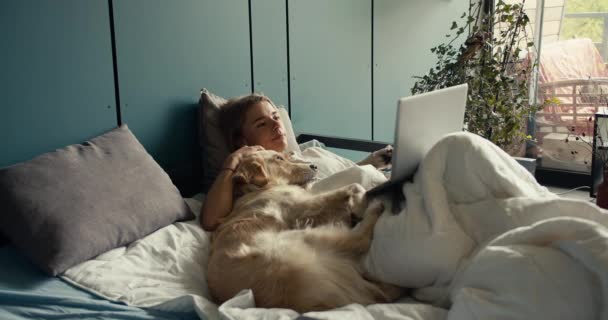 ブロンドの女の子が犬とベッドに横たわり 朝のラップトップで映画を見ている ペットとの休日 — ストック動画