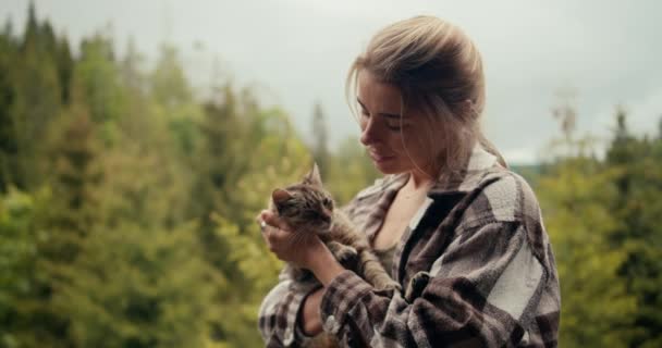 クローズアップショット 幸せなブロンドの少女は彼女の腕に猫を保持し 山の複雑な森の背景に対してバルコニーに彼女をストローク — ストック動画