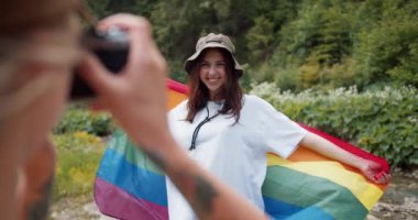 Beyaz bir tişörtle yeşil bir ormanın arka planına yaslanmış bir LGBT bayrağıyla duran arkadaşının fotoğrafını çeken bir kızın görüntüsü..