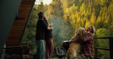 A group of friends on a picnic overlooking the mountains and coniferous forest. A brunette guy is hugging a brunette girl, and a blond man is stroking a large light-colored dog.