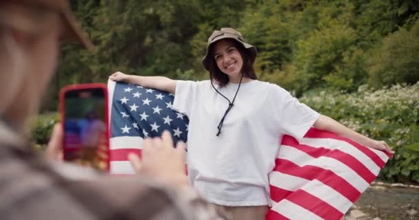 一个金发女郎在森林的背景下调整了一下 说如何站起来 让一个身穿白衬衫 挂着美国国旗的黑发姑娘拍一张更好的照片 — 图库视频影像