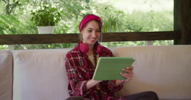 赤いヘッドフォンと赤い包帯を持つ赤い再生シャツのブロンドの女の子が座っており 自然の中でソファの上に緑のタブレット上で動作します — ストック動画