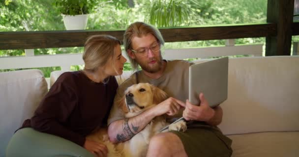 ガラスの髭を生やした金髪の女の子と金髪の男がラップトップで何かを見ている 犬が隣に座っている 自然の中でソファの上でのレジャー — ストック動画