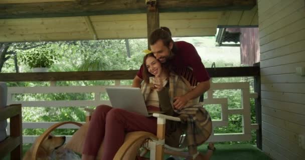 一个穿着红色T恤的黑发男人围住一个坐在扶手椅上的女孩 看着笔记本电脑屏幕 在森林的背景下喝茶 对儿童的照料的表现 — 图库视频影像