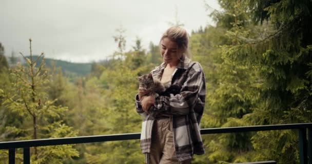 快乐的金发姑娘在一座俯瞰高山针叶林的乡间房子的阳台上爱抚她的猫 — 图库视频影像