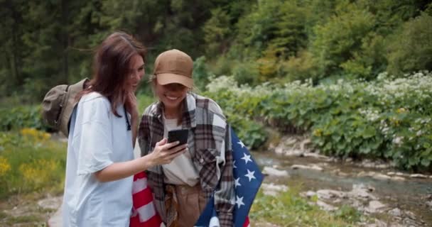 ブルネットの少女は アメリカの国旗を持った友人の写真を撮り 彼らは写真を見ています 緑の森と山の川の背景に対する写真 — ストック動画