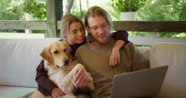 一个金发女郎和一个戴眼镜的金发女郎正在看笔记本电脑上有趣的东西 他们的狗就坐在他们旁边笑着 在大自然的凉亭里消闲 — 图库视频影像