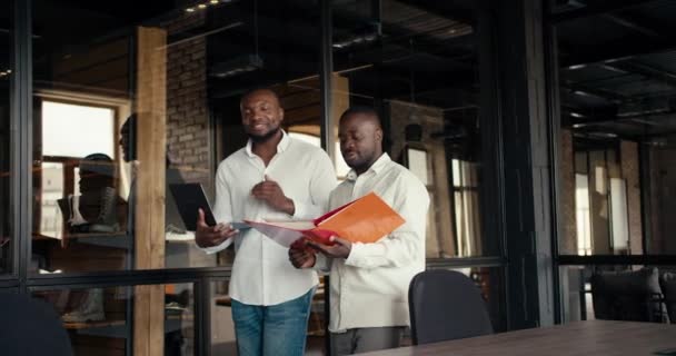 白いシャツを着た2人の黒人のビジネスマンがロフトスタイルのオフィスを歩き回り ラップトップと書類付きのフォルダを使用して新しいプロジェクトを議論しています — ストック動画