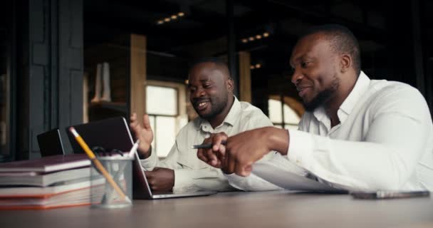 两名白衬衫黑皮肤的办公室职员坐着讨论笔记本电脑上的实际问题 在办公室的纸张和手提电脑上解决紧急问题 — 图库视频影像