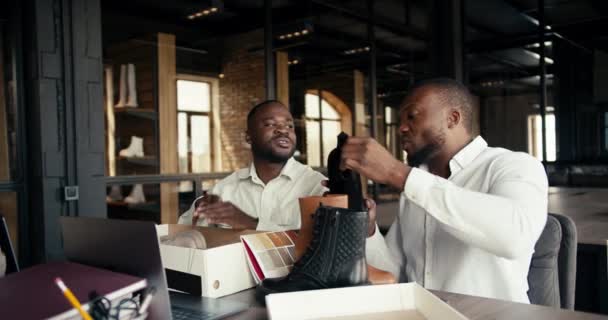 两名身穿白衬衫的黑皮肤商人正在讨论一种新的颜色和鞋子的设计 并以摆在桌上和盒子里的鞋子为例 — 图库视频影像