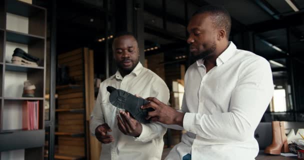 靴工場の2人のデザイナーマネージャーが新しい靴のバッチについて話しています 白いシャツの2人の黒人男性が工場でオフィスで新しいスタイルの靴を議論しています — ストック動画