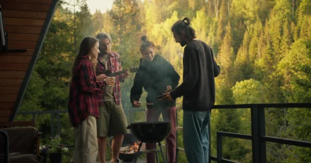 一群朋友在乡间小屋的阳台上 在一片绿色针叶林的背景下 正在悠闲地烧烤着肉 这家伙弹吉他 两个人一起工作 — 图库视频影像