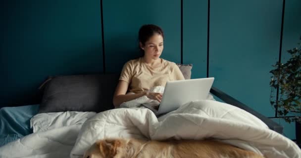 一个黑发女孩躺在床上 在笔记本电脑上工作 在家里的青绿色墙壁后面笑着 从家里远程工作快乐 — 图库视频影像