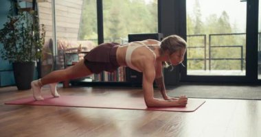 Beyaz bluzlu sarışın bir kız yeşil ormana bakan endüstriyel bir evde özel bir paspas üzerinde dinamik bir tahta egzersizi yapıyor. Evde sağlık, beden ve zihin güçlendirme.