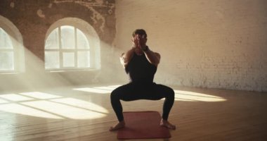 Sporcular siyah formalı sıkı sporcular çömelmiş ve kollarını sallıyor. Tuğla duvarlı güneşli bir spor salonunda sabah sporu..