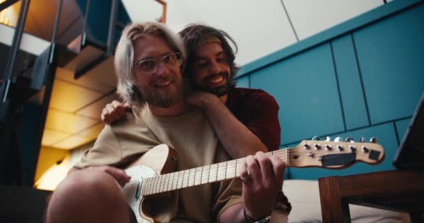 2人の友人が一緒に時間を過ごす男ブルネットは居心地の良い部屋で電気ギターを演奏するメガネでブロンドを抱きしめる — ストック動画